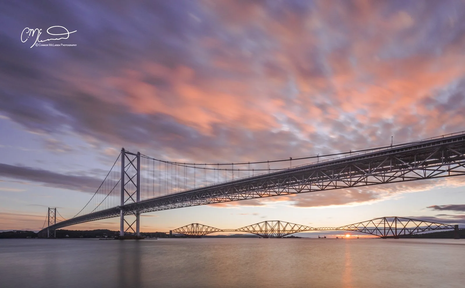 Scottish Lowlands, Scotland, Forth bridge Edinburgh. Sunset over a large suspension and cantilever bridge spanning over water, with pink and purple clouds in the sky.