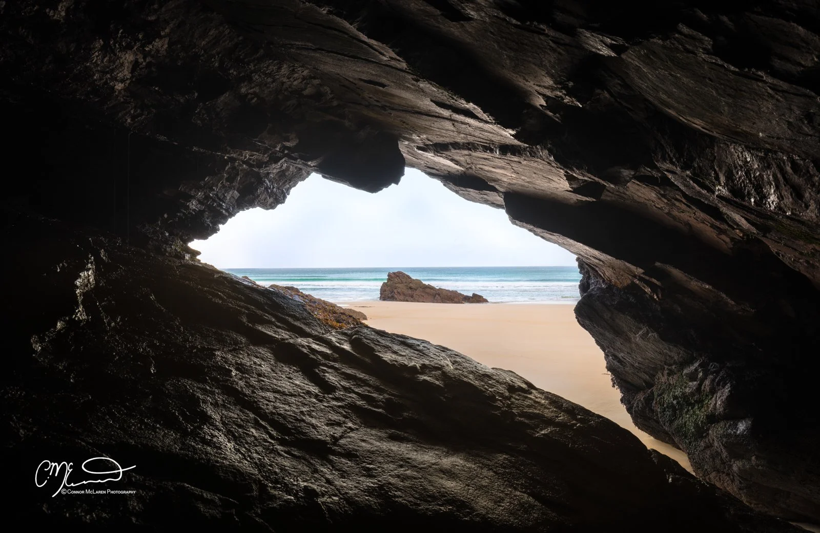 Scotland, Lewis Harris & Outer Hebrides, Tolsta. View through a rocky cave opening to a sandy beach, ocean waves, and a large rock on the shore.