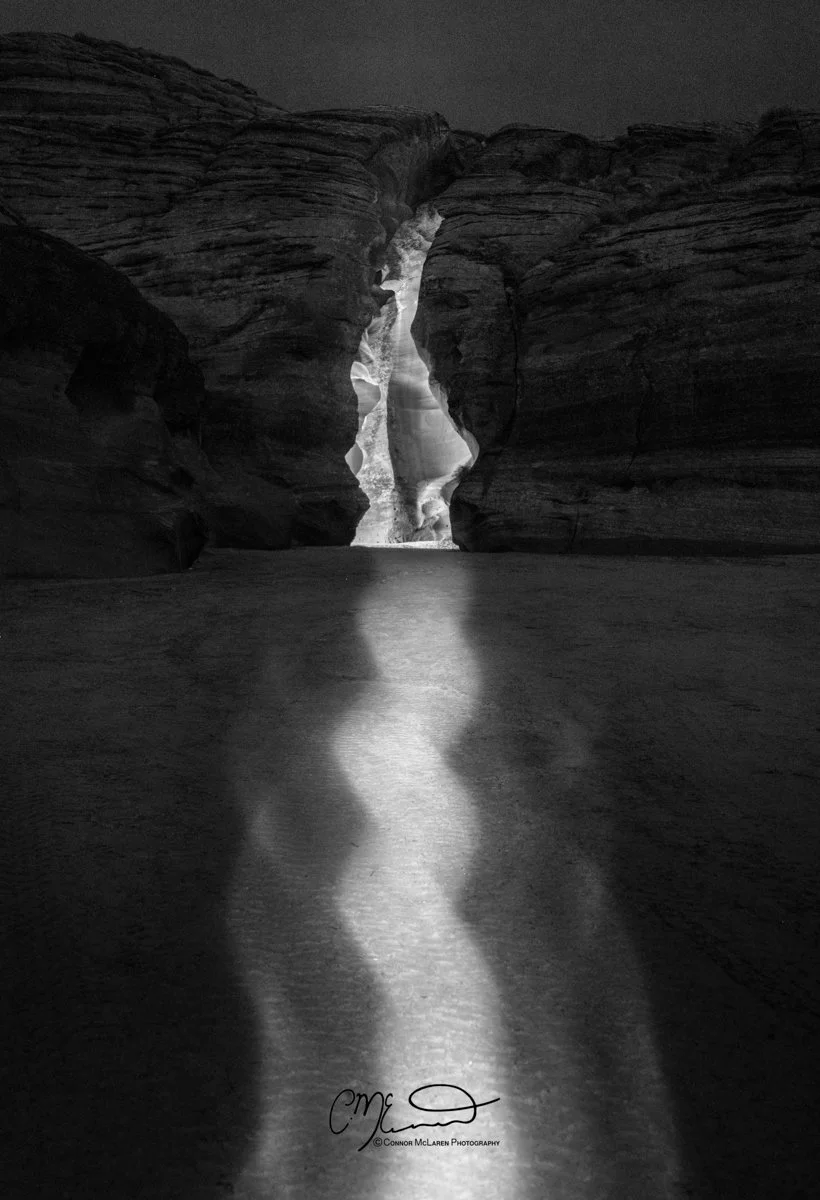 USA Antelope Canyon Arizona. A black and white photo of a narrow canyon with a beam of light shining down onto a flat surface, possibly a desert floor, with towering rock walls on either side.