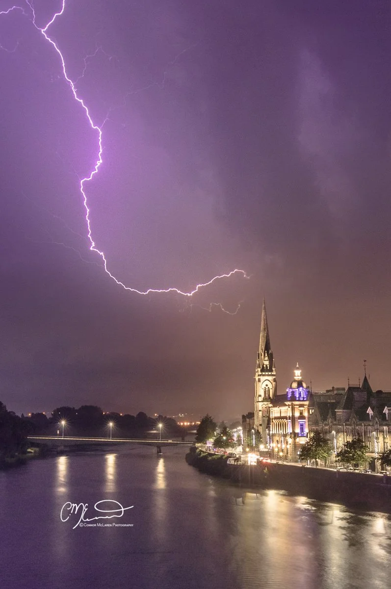 Perth Scotland Lightning storm. Lightning storm over a cityscape at night, reflecting on a river, with historic buildings and a church with a steeple, lit with purple and white lights.