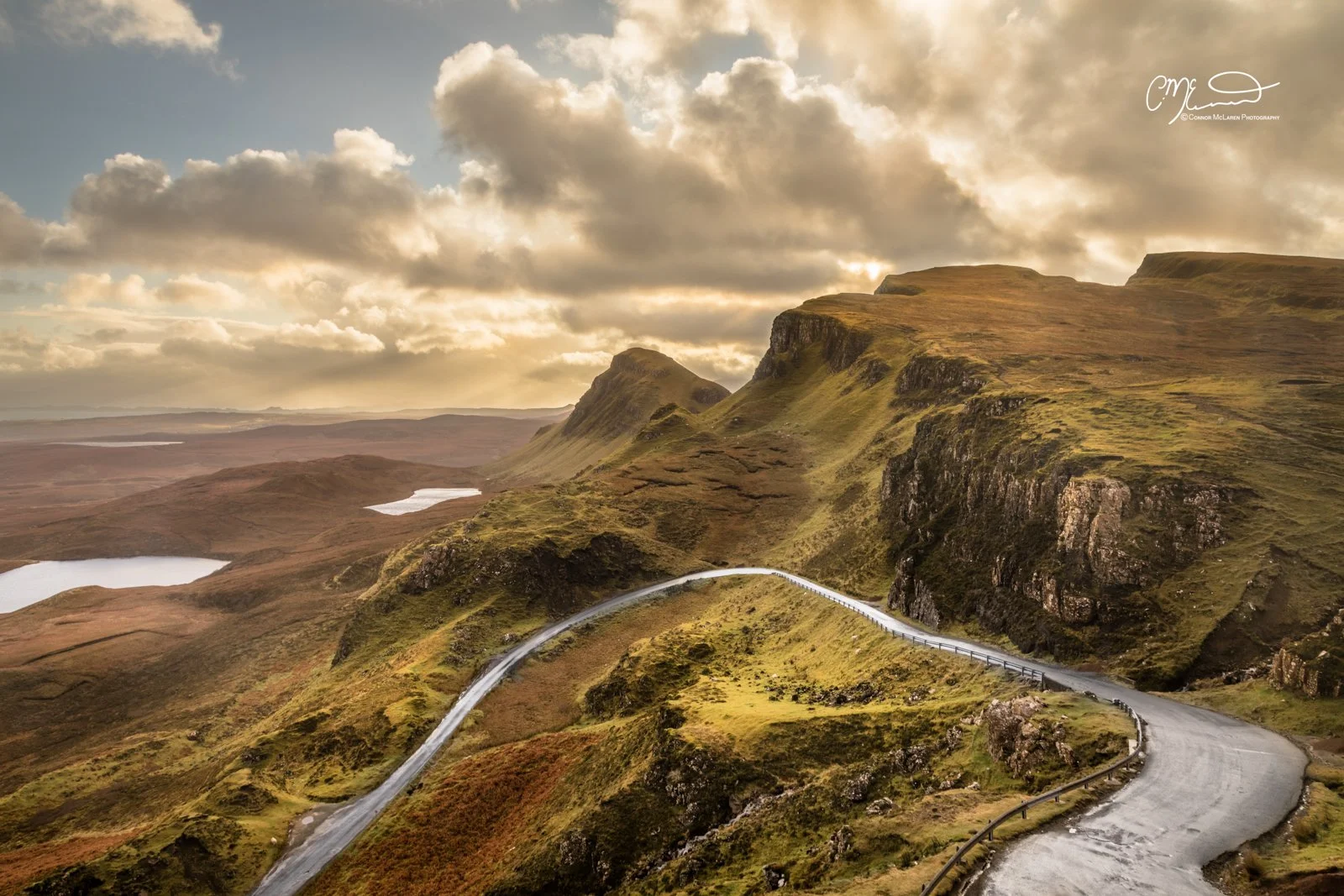Scotland, Isle of Skye, Quiraing. A winding mountain road through a hilly landscape with grassy slopes and rocky cliffs under a cloudy sky during sunset.
