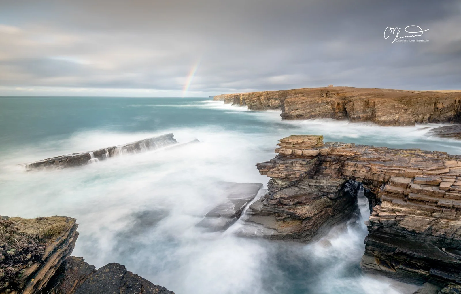 Yesnaby, rainbow, Orkney, Scotland. Cliffs and rocky shoreline along a turbulent ocean, with a faint rainbow in a cloudy sky.