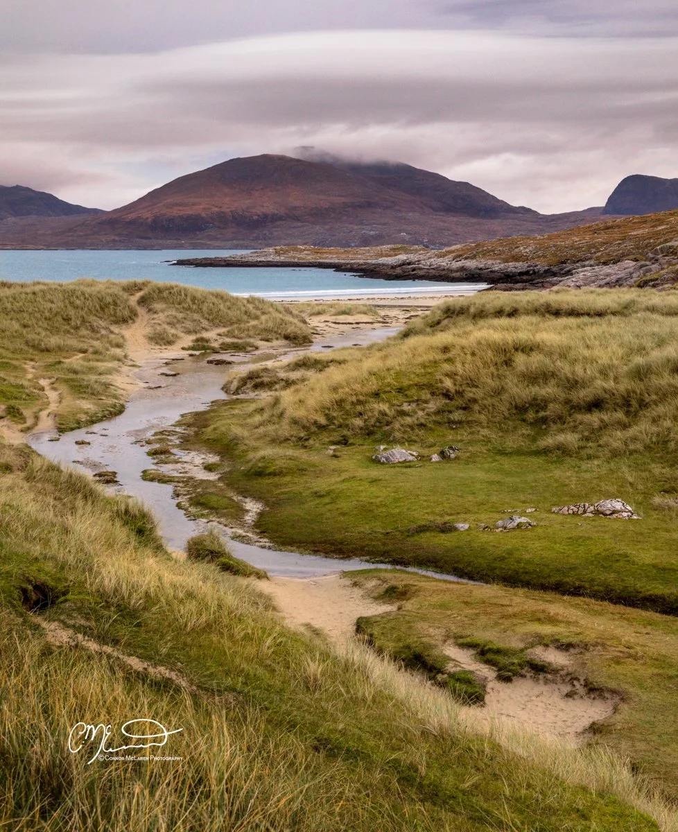 Scotland, Lewis Harris & Outer Hebrides, Luskentyre Beach. Scenic landscape featuring grassy dunes, a small stream, sandy paths, a body of water, and mountains in the background under a cloudy sky.