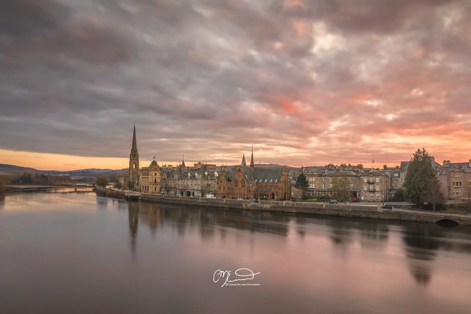Perth Scotland, Scottish central highlands. A cityscape by a river during sunset with historic buildings and church steeples under a partly cloudy sky.
