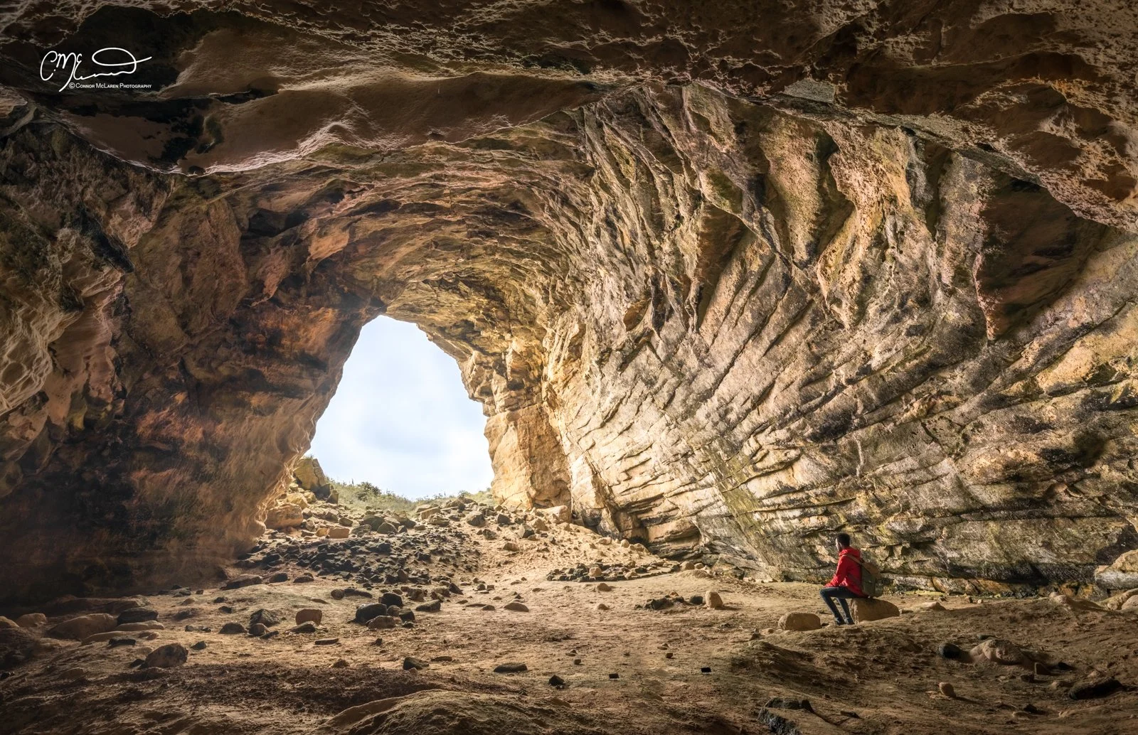 Covesea Caves, North East Scotland. A person in a red jacket sitting on a rock inside a large cave with layered rock formations and an opening revealing the sky outside.