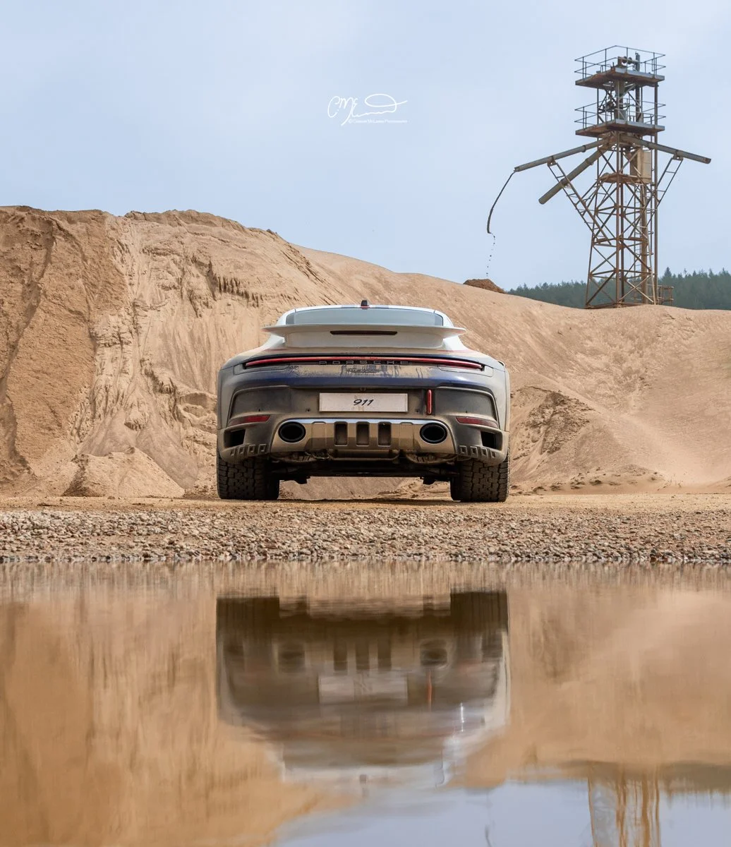 A Porsche 911 Dakar Roughroads sports car parked on a gravel surface in front of a large dirt mound, with a reflective puddle of water in the foreground and a metal tower structure with a pipe or arm extending from it in the background.