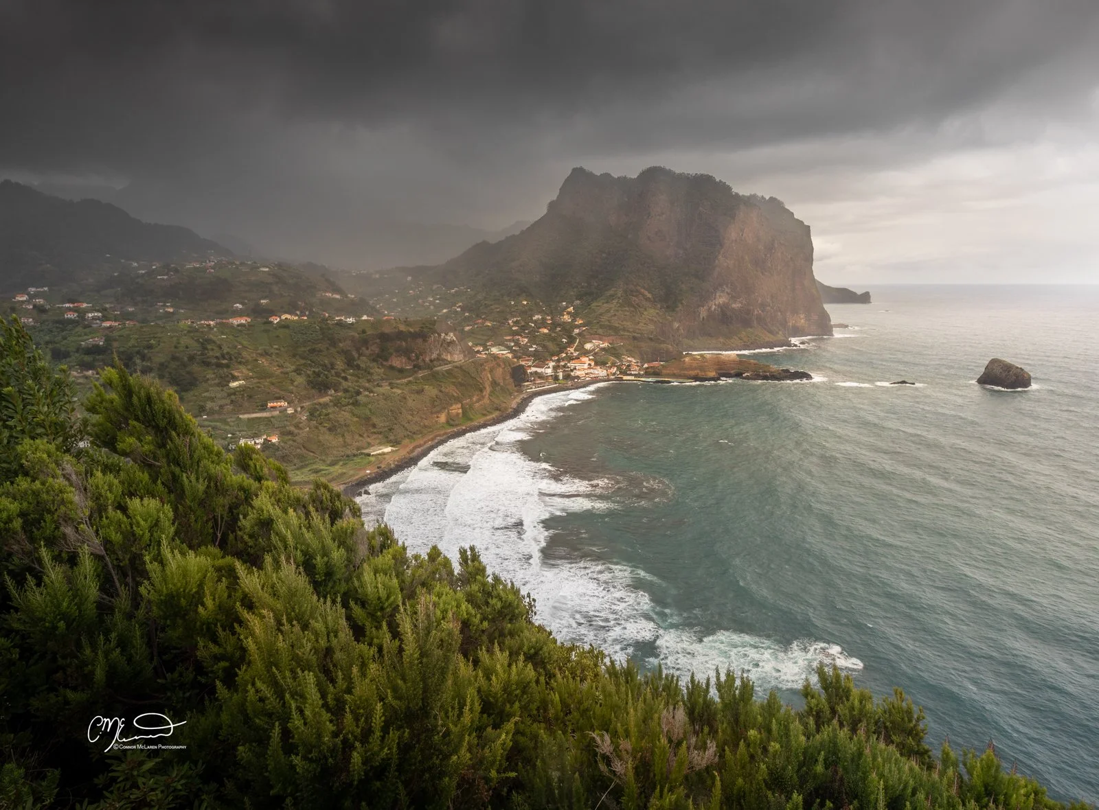 Volcanic rock, porto da cruz, Madeira Portugal. Waterfront view of a rocky coastline with waves, a small town at the base of green mountains, and dark storm clouds above.