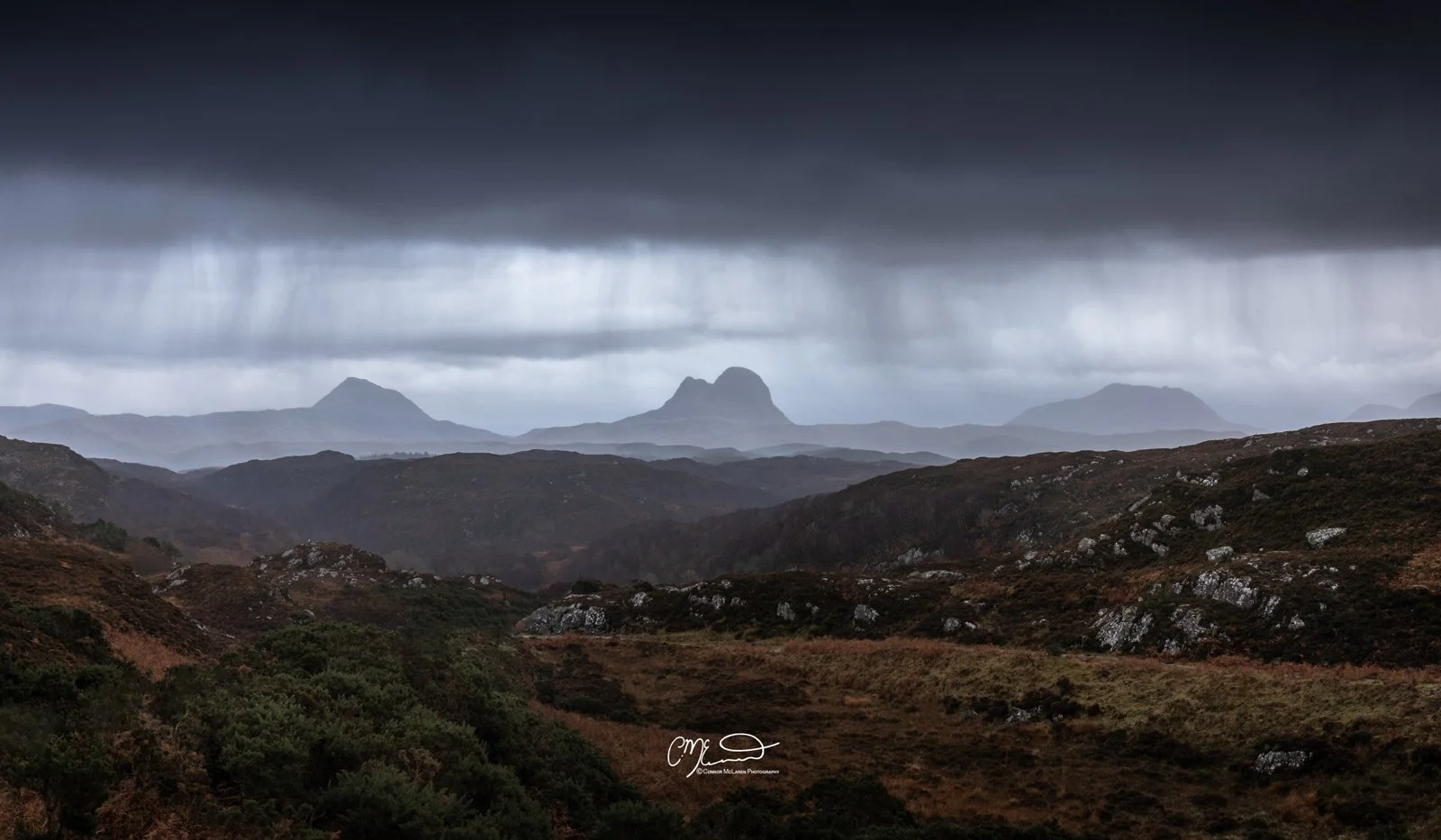 Scotland, north coast 500, NC500, Assynt. A dramatic landscape with dark, stormy clouds over a series of mountains and rolling hills in the distance, with mist and fog creating a moody atmosphere.