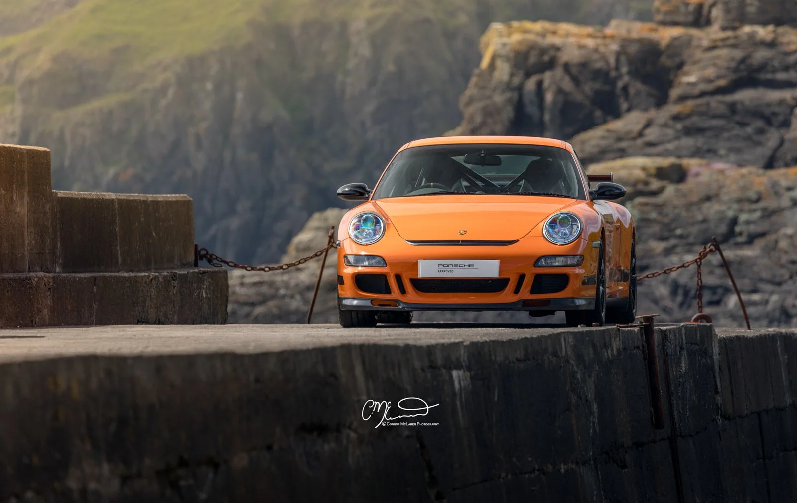 Porsche 911 GT3 RS, 997. Orange Porsche sports car parked near stone barriers with a rugged mountain landscape background.