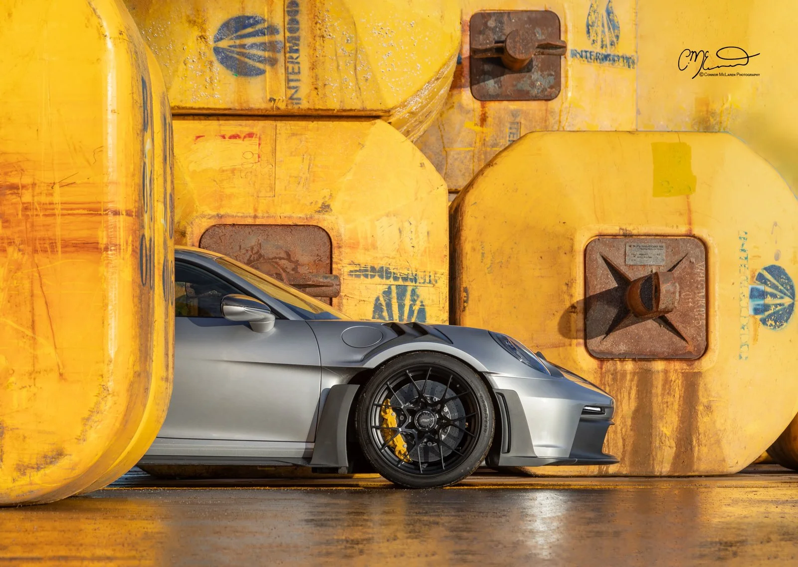 A silver Porsche 911 GT3 RS sports car with black wheels and yellow brake calipers is parked in front of a large yellow industrial storage tank.