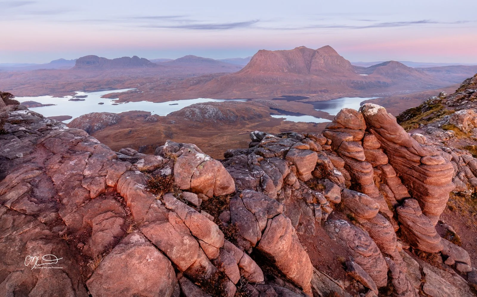 Scotland, north coast 500, NC500, Assynt. Stac Pollaidh. Rocky viewpoint overlooking a scenic landscape with lakes, rolling hills, and distant mountains at sunset in a rugged, natural setting.