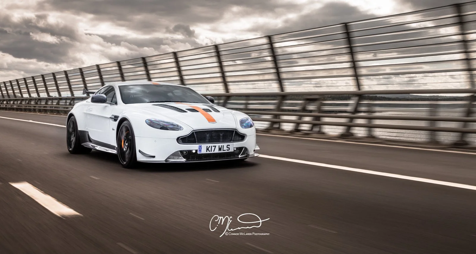 White Aston Martin V12 Vantage amr sports car with orange stripe driving on a highway overpass, cloudy sky background.