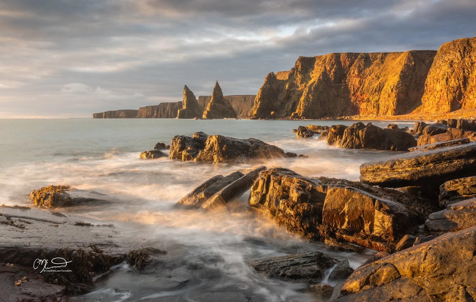 Bucket list locations, Scotland, north coast 500, duncansby head stacks. . Sunset at a rugged rocky coastline with cliffs and sea stacks in the distance, gentle waves washing over rocks in the foreground.