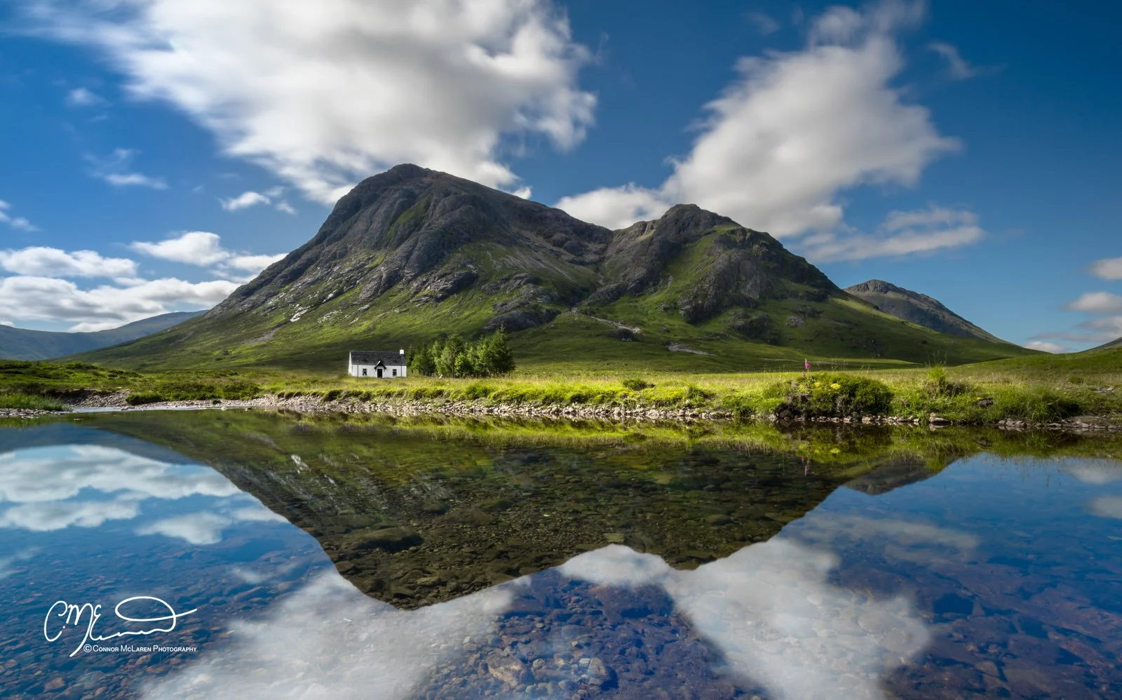 Glen Coe and Glen Etive, Western Highlands, Scotland. A scenic landscape of a mountain with lush green slopes reflected in a calm lake, with a small white house and trees nearby under a partly cloudy sky.