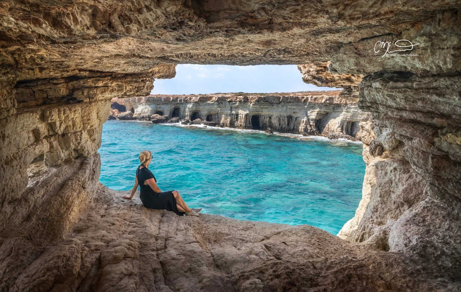 Ayia napa, Cyprus. A woman sitting on a rocky ledge inside a cave opening, overlooking blue ocean water and rocky cliffs.