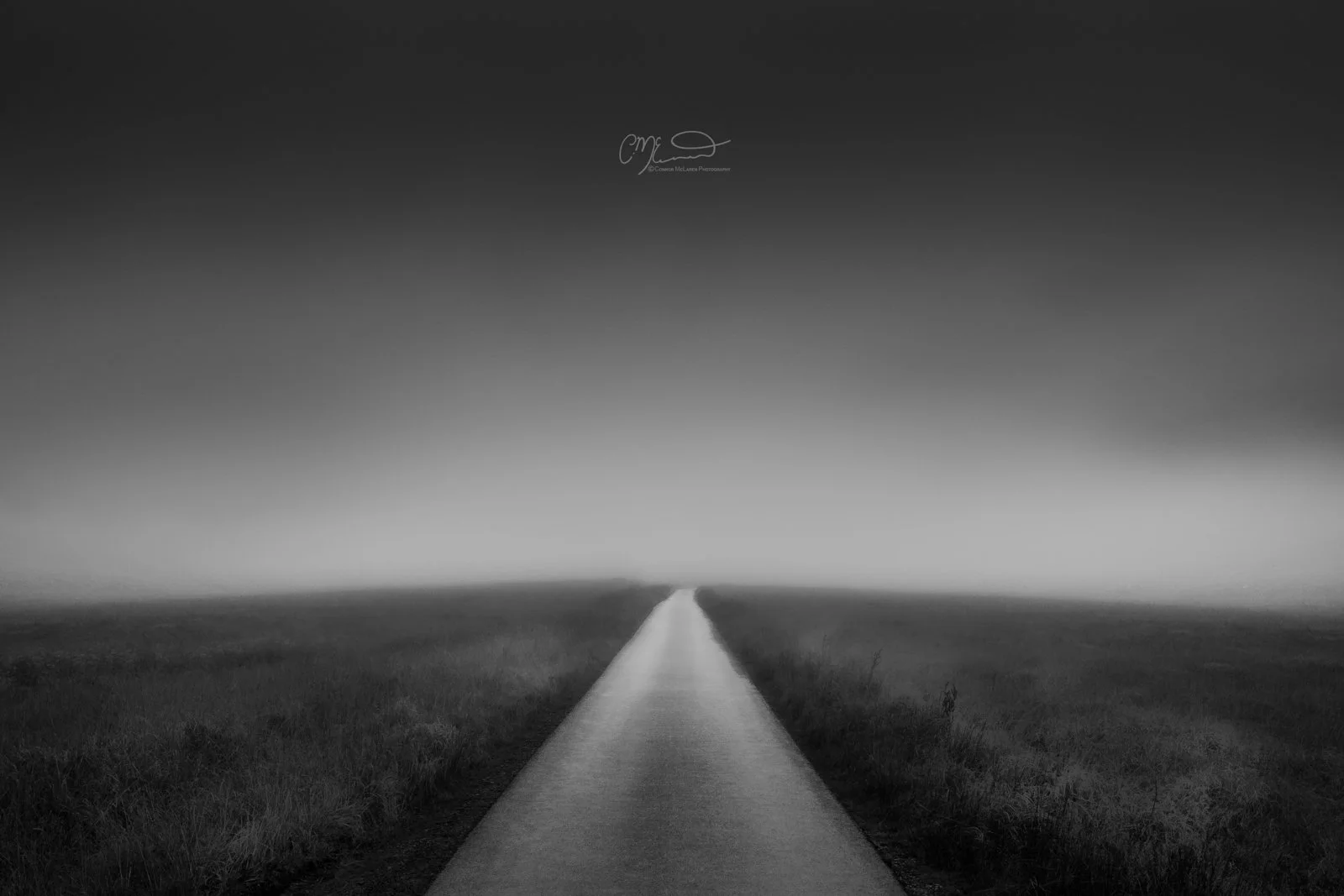 Scotland, Glen Etive, Skyfall Road. A long, straight road extending into the distance surrounded by flat open fields under a dark, cloudy sky in black and white.