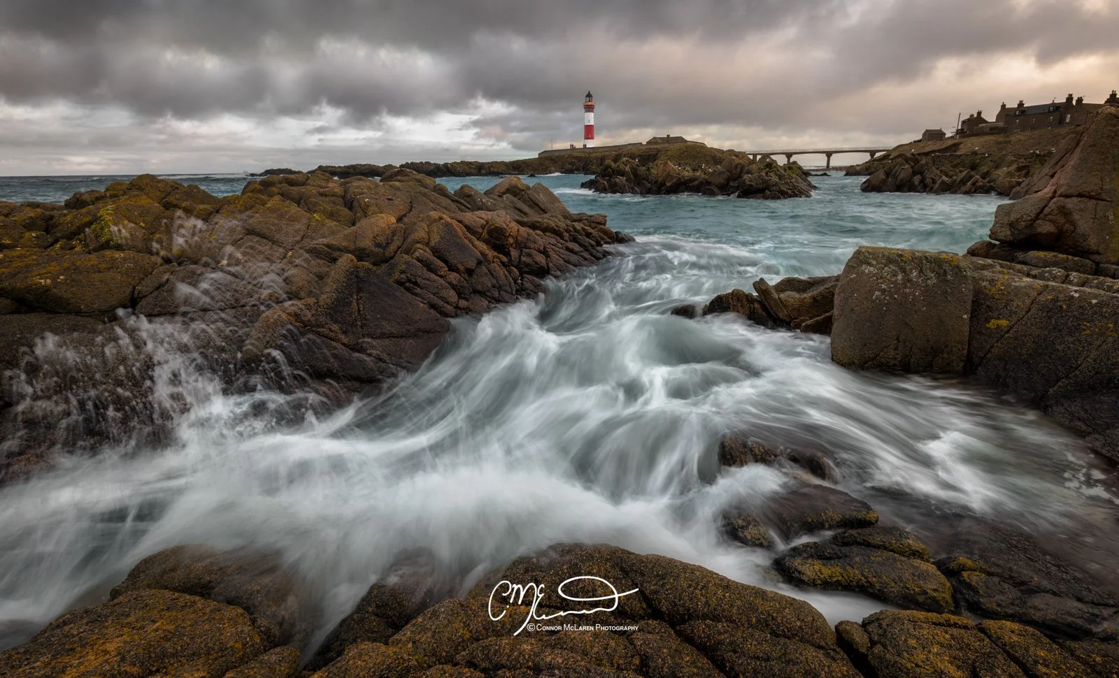 boddam lighthouse, storm, North East Scotland. A rough shoreline with large rocks, turbulent water, a lighthouse in the distance, and houses on the cliffs under cloudy skies.