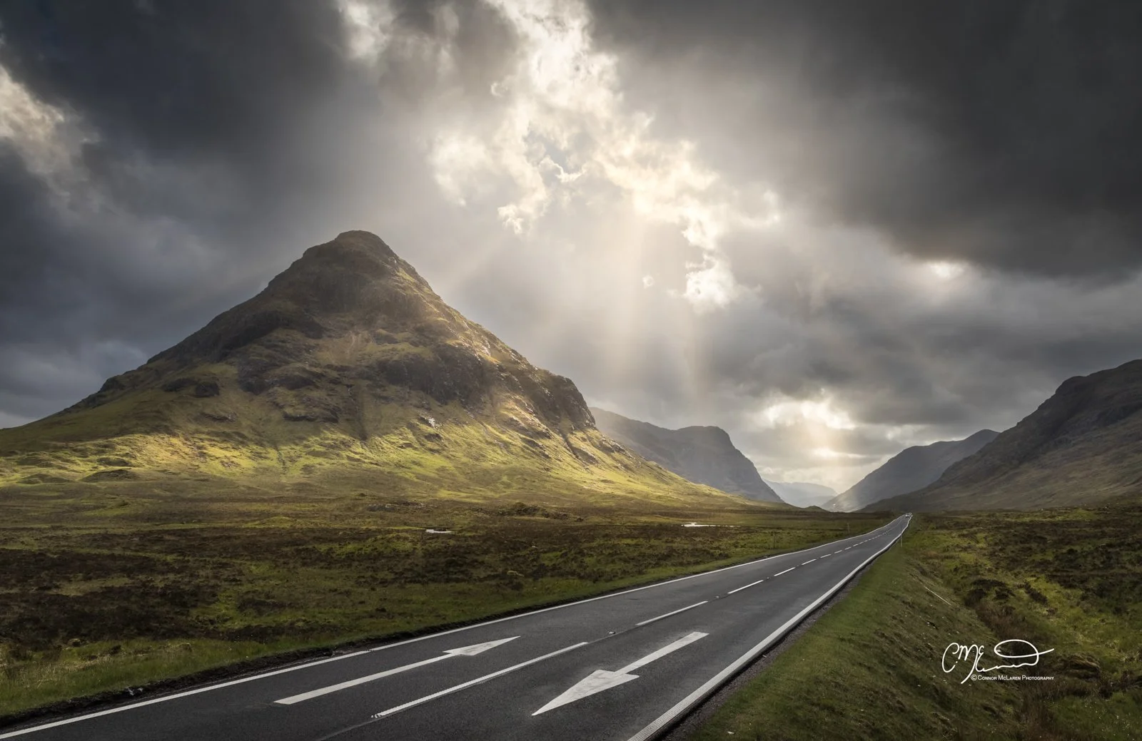 Glen Coe and Glen Etive, Western Highlands, Scotland. A long winding road cuts through a green valley flanked by mountain peaks under a dramatic cloudy sky with sunlight breaking through.