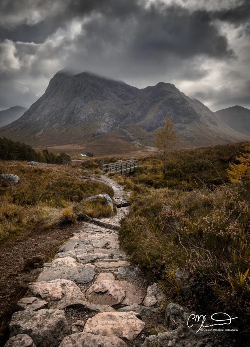 Glen Coe and Glen Etive, Western Highlands, Scotland. A rocky trail winding through a grassy landscape with a wooden bridge, leading towards a mountain under dark, cloudy skies.