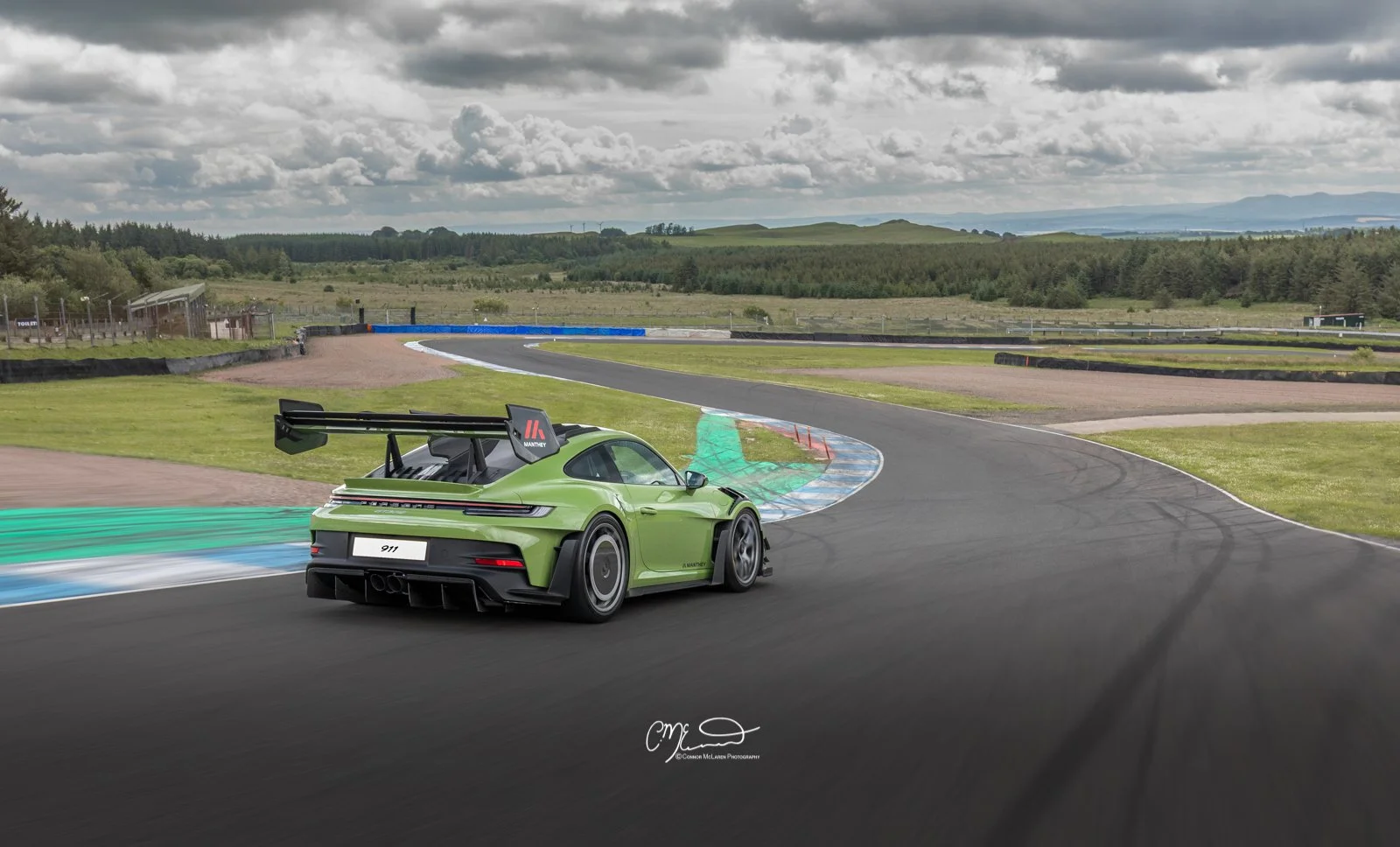 A green Porsche 911 race car on a racing track, taking a turn with clouds overhead, countryside background, and safety barriers along the track.