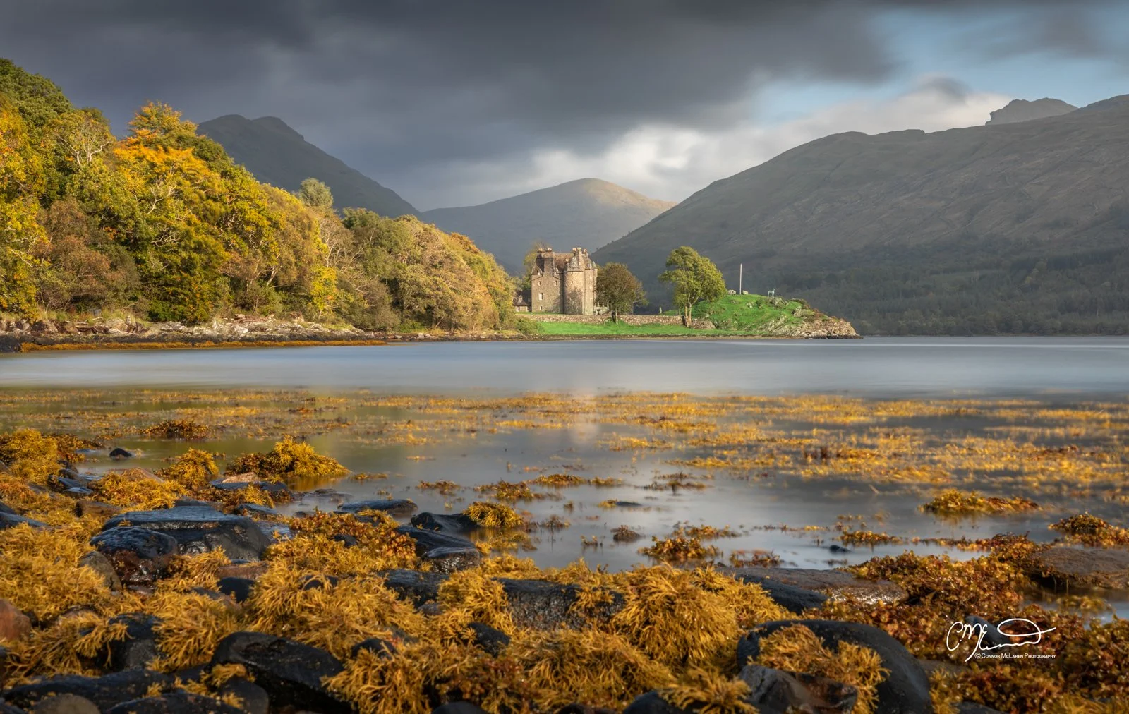 Scottish castle, Western Highlands, Scotland. A scenic landscape of a lake with rocky shoreline and seaweed in the foreground, a castle and trees on the opposite shore, and mountains with dark clouds in the background.