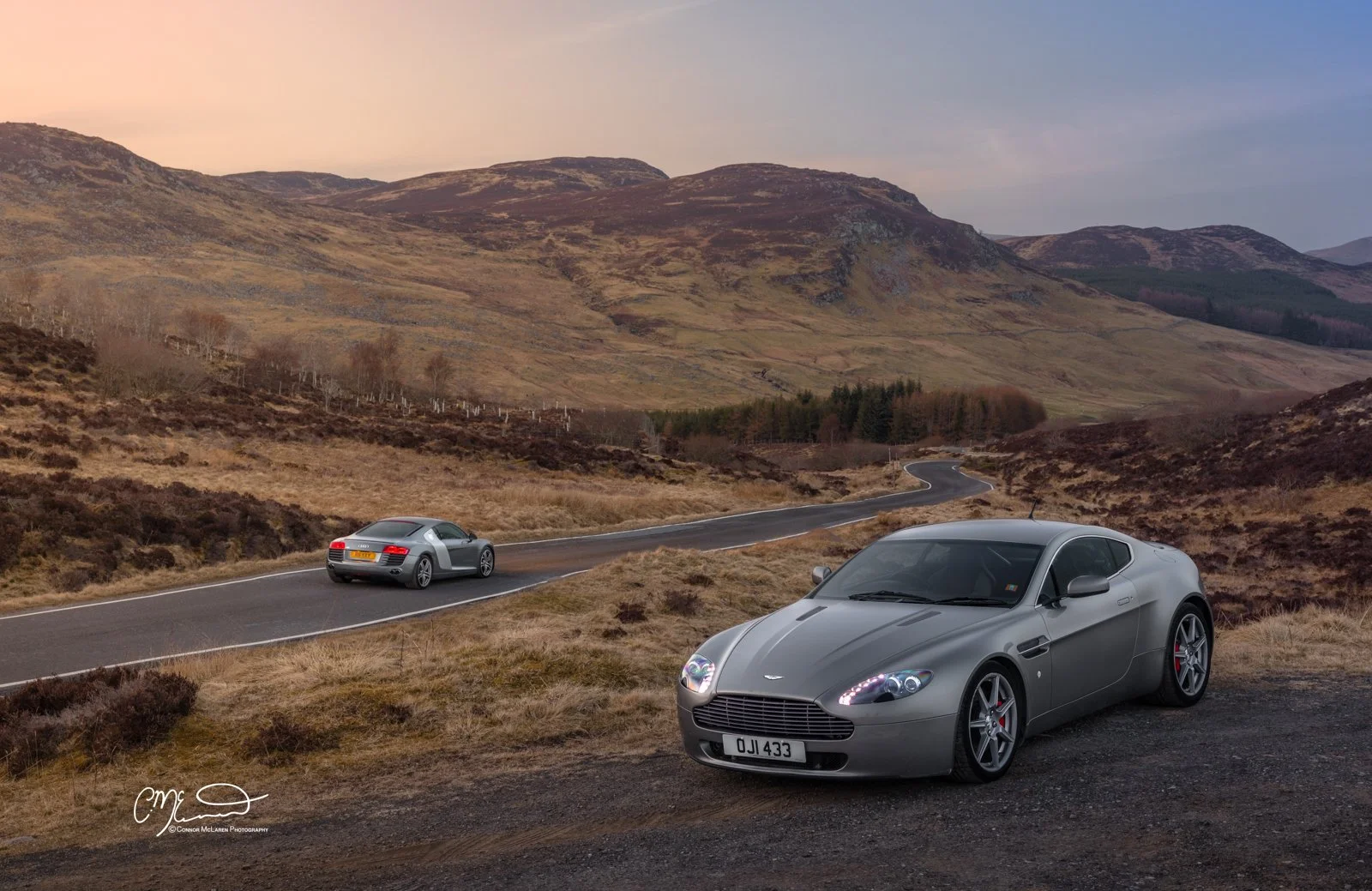 Two grey sports cars parked on a mountain road with a landscape of hills and mountains in the background during sunset.