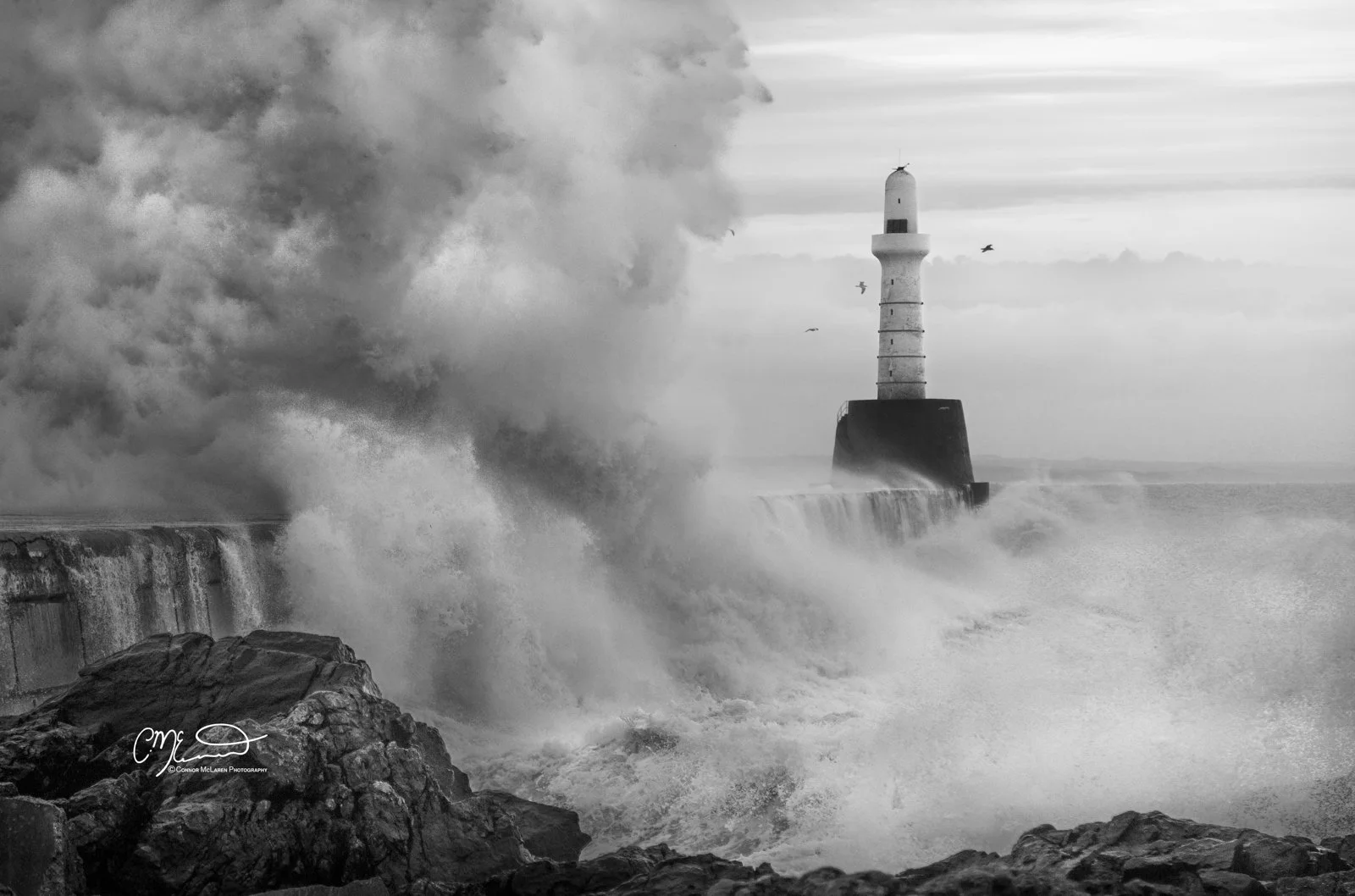 Scotland, Aberdeen Lighthouse, waves. Black and white photo of a lighthouse on a breakwater with large waves crashing against it and the rocks in the foreground.