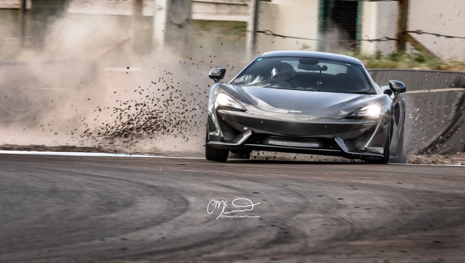 A sleek, gray McLaren 570s race car speeding on a track, kicking up dirt and debris as it takes a turn.