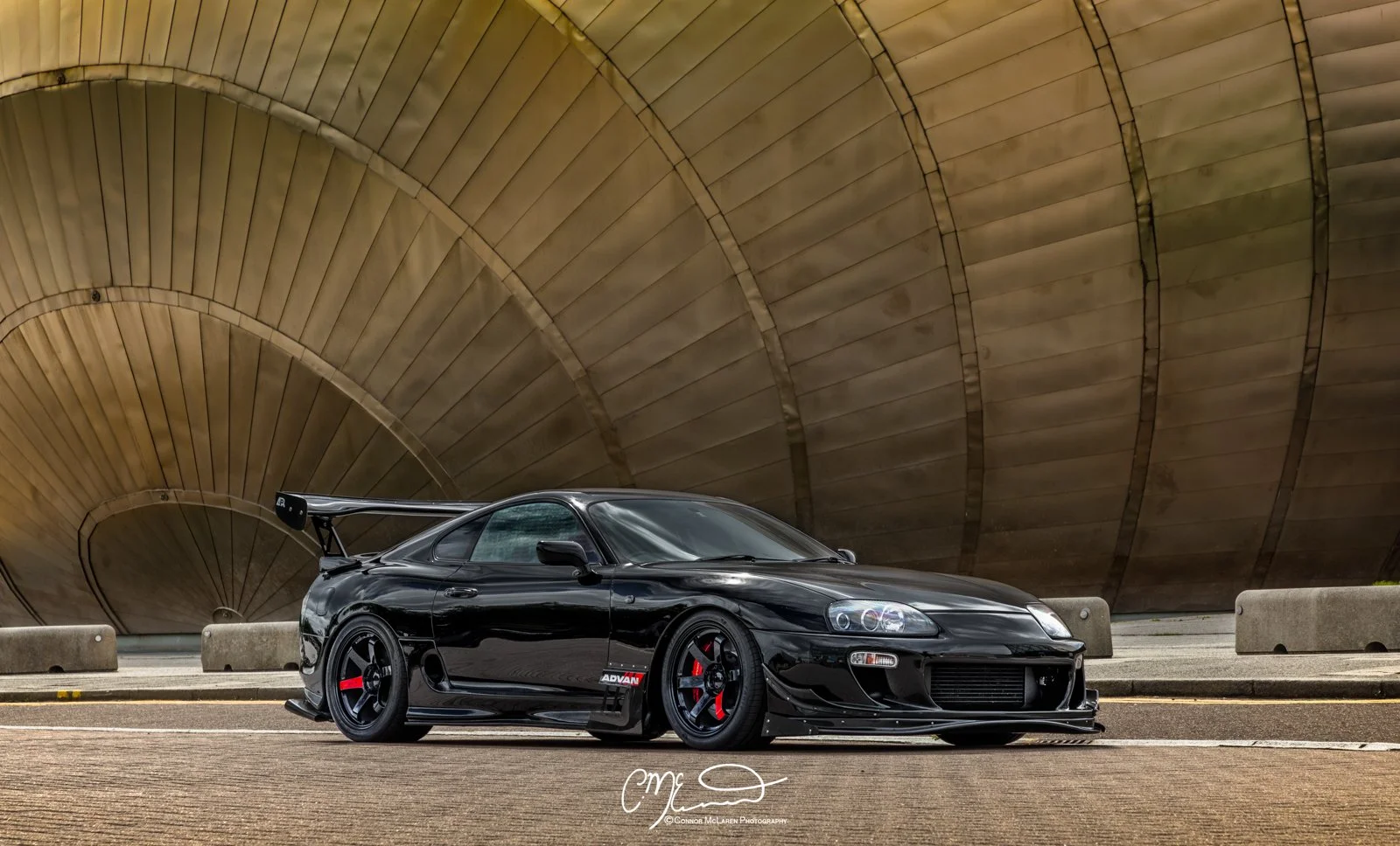 Custom Modified Toyota Supra 2JZ. A black sports car with a large rear spoiler parked on a road in front of an abstract, curved, metallic structure.