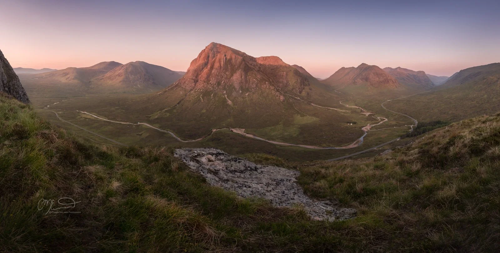 Glen Coe and Glen Etive, Western Highlands, Scotland. A scenic view of a valley with winding roads and mountain peaks at sunrise or sunset. The central mountain is illuminated with pinkish light, with a clear sky above.