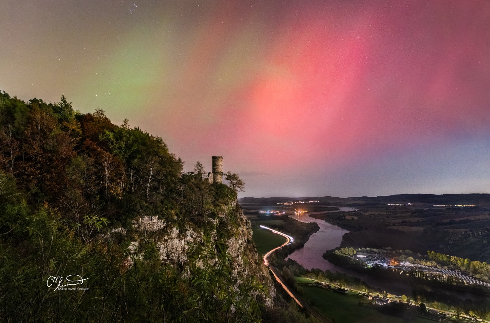 Northern Lights Perthshire, Kinnoull Hill, PeNighttime landscape showing a river winding through a valley, with a forested cliff on the left side featuring an old stone tower, and the sky illuminated with colorful auroras in shades of green and pink.