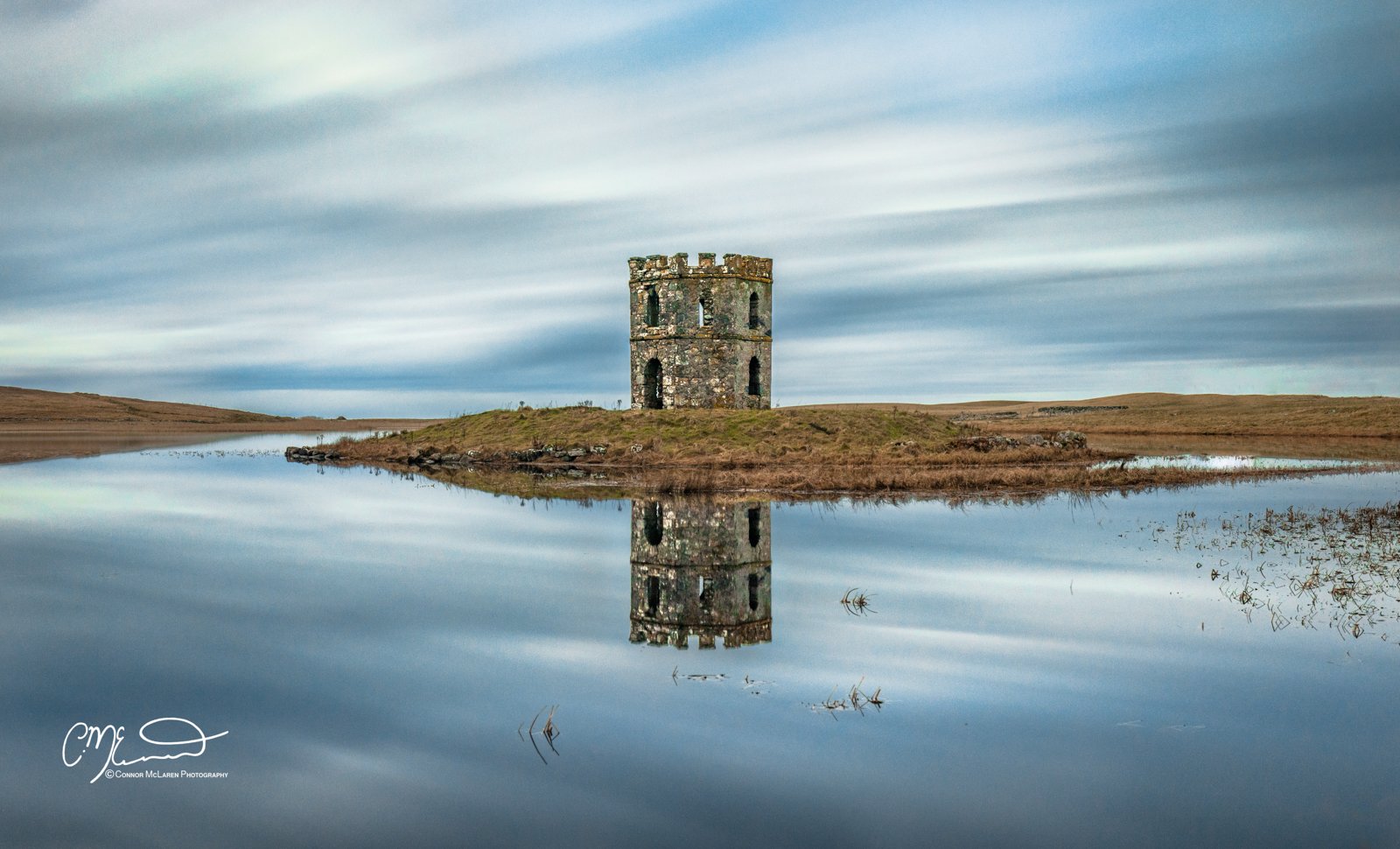 Scotland, Lewis Harris & Outer Hebrides, north Uist. Old stone tower on a small island, reflected in calm water with grassy surroundings and a cloudy sky.