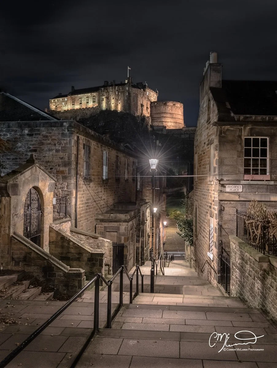 Edinburgh Castle. Nighttime view of a steep stone staircase leading down to a quiet street, with old stone buildings on either side, street lamps illuminating the scene, and a castle on a hill illuminated in the background.