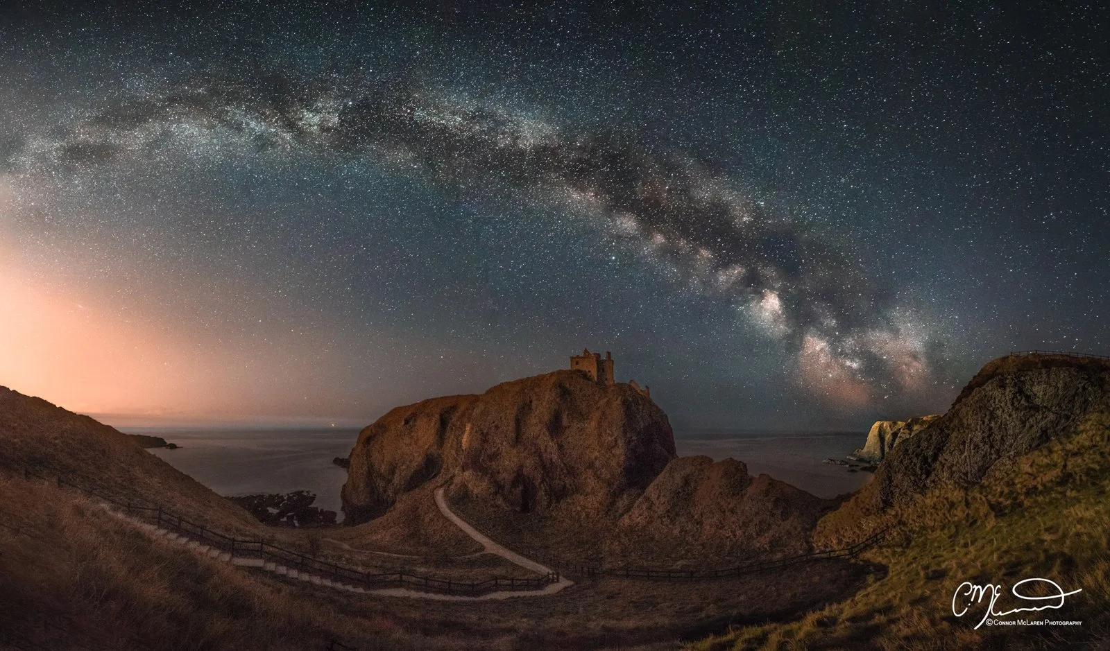 Dunnottar Castle, North East Scotland. Nighttime landscape of a rocky coastline with a castle on a large rock formation, under a starry sky with the Milky Way galaxy visible.