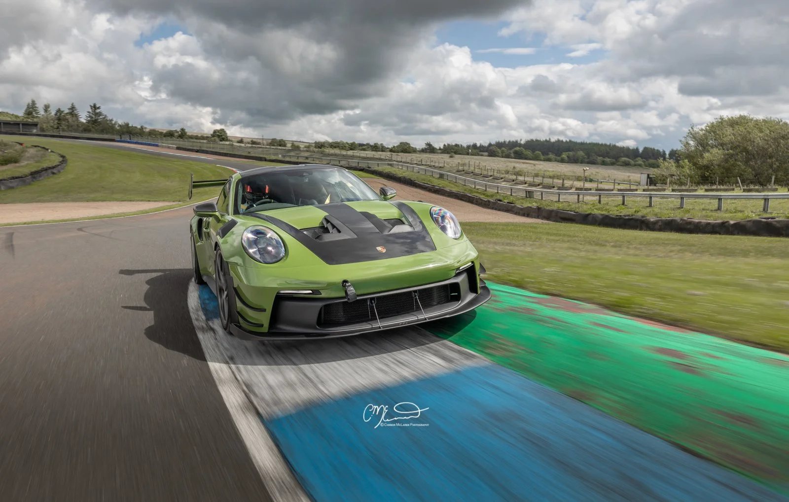 Porsche 911 GT3 RS with Manthey Racing kit in PTS Olive Green, Knockhill racing. Amazing photograph, wallpaper. A green Porsche race car on a track with colorful markings during the day with a cloudy sky.