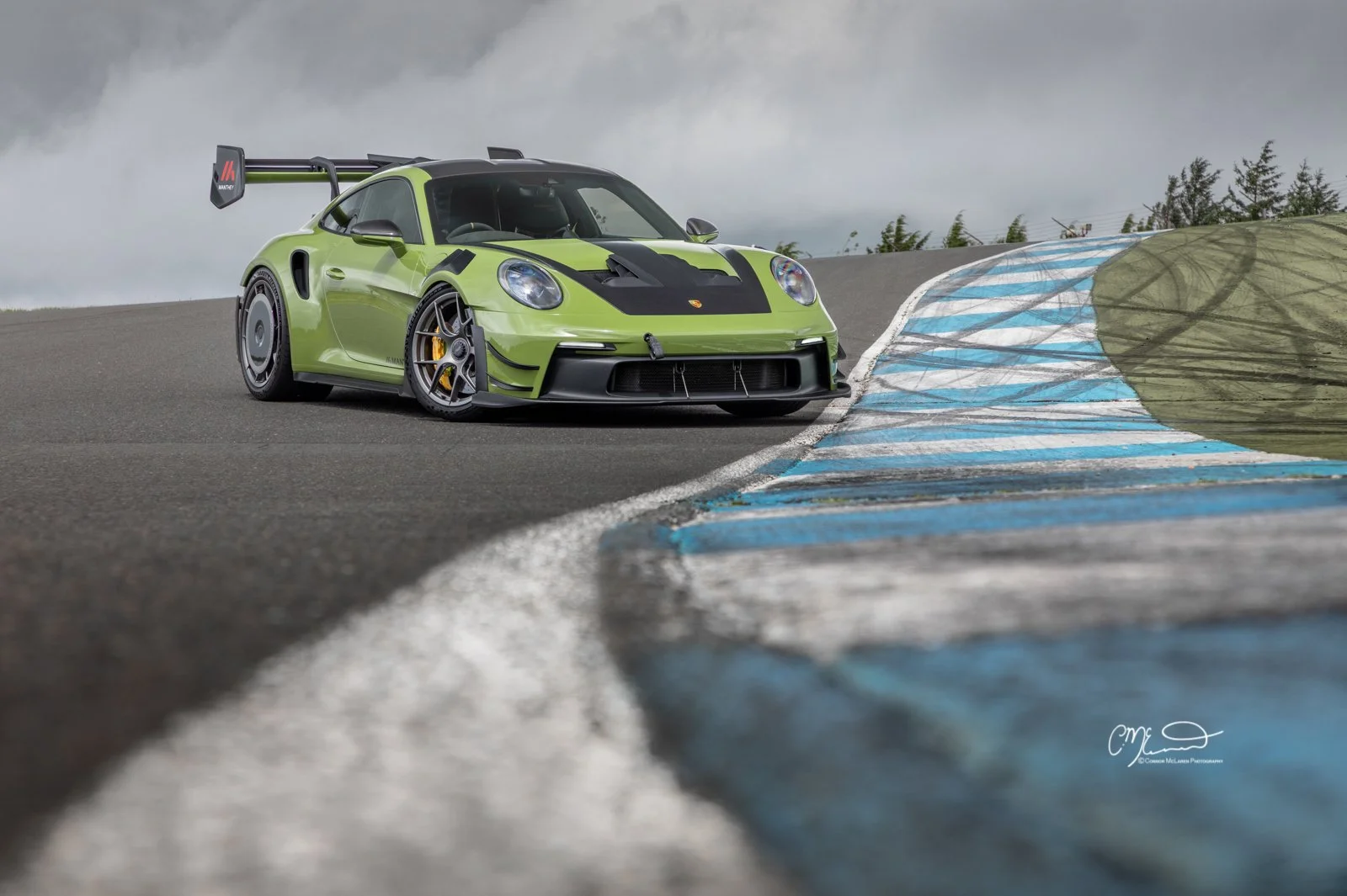 Porsche 911 GT3 RS with Manthey Racing kit in PTS Olive Green, Knockhill racing. Amazing photograph, wallpaper. A green race car on a track with blue and white painted curbs and cloudy sky in the background.