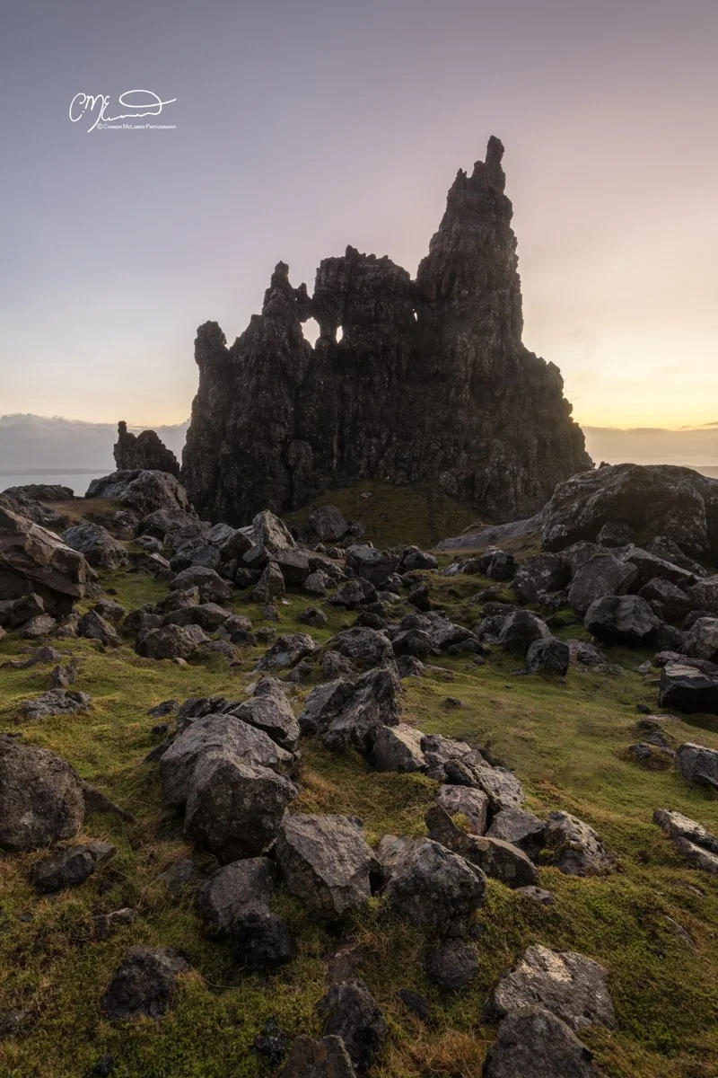 Scotland, Isle of Skye, Old Man of Storr. A tall, rugged rock formation standing amidst a rocky landscape with patches of green moss and grass, against a sunset sky.