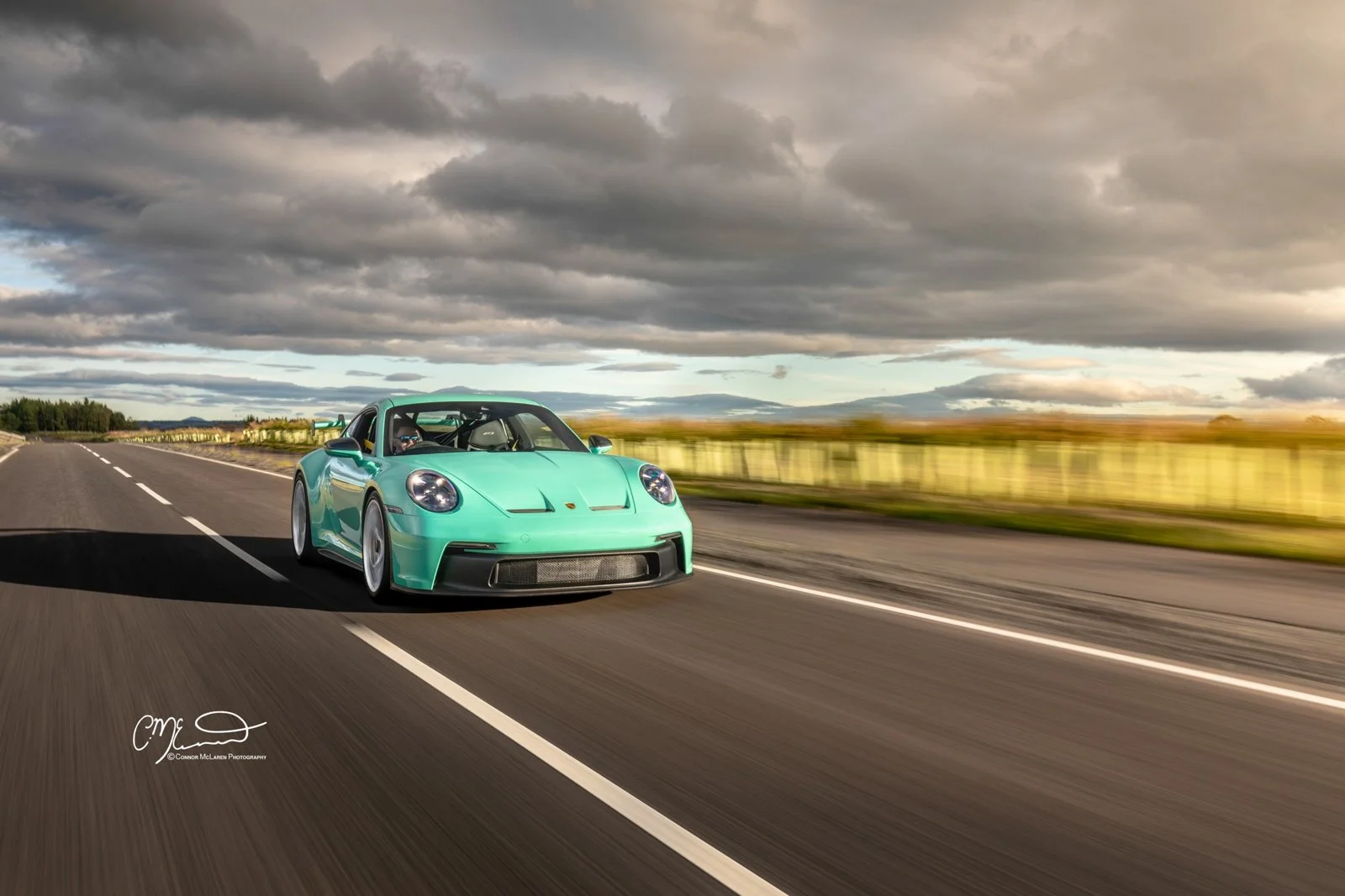 Mint Porsche 911 GT3 992. A mint green sports car driving on a two-lane road with a blurred landscape of fields and hills in the background under a cloudy sky.