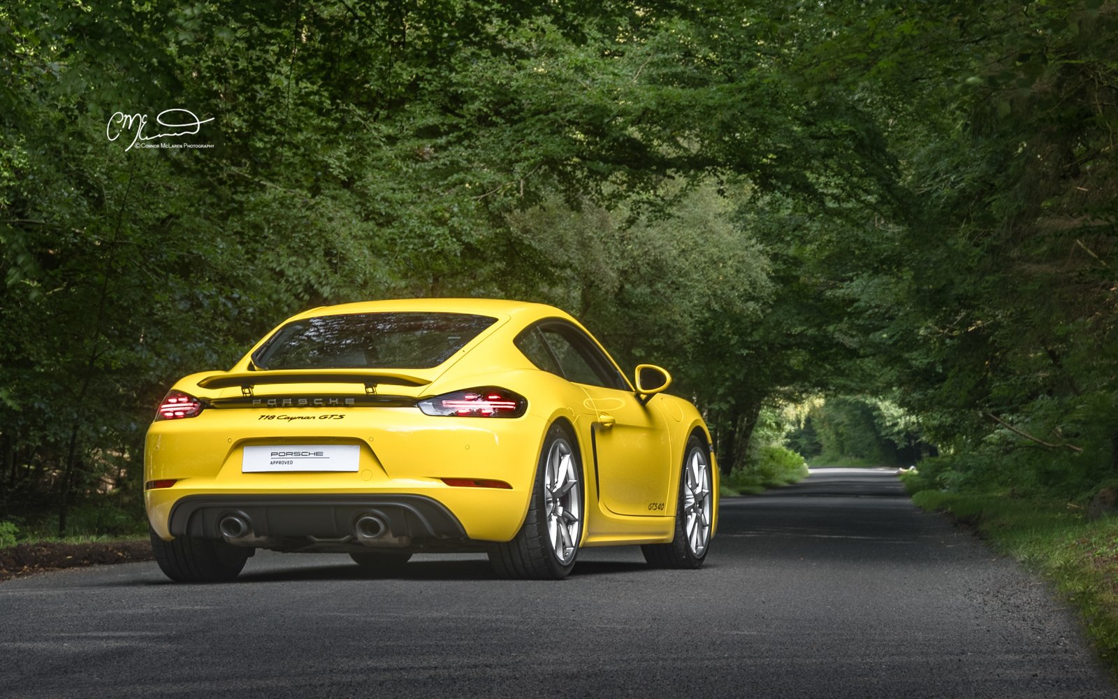 A yellow Porsche 718 Cayman GTS parked on a tree-lined road.