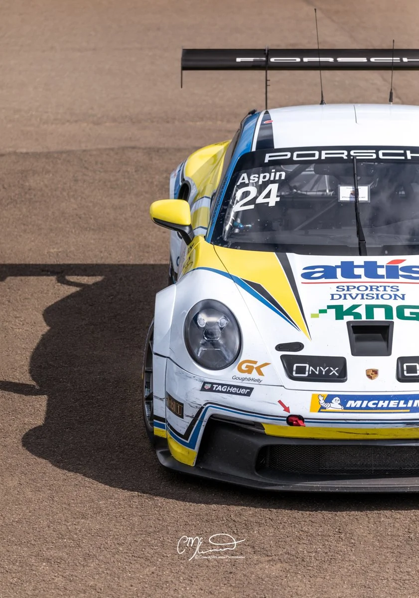Porsche 911 GT3 Cup car. Racecar. Close-up of a race car with sponsor logos, yellow and white body, on a tan racing surface, with shadows cast on the ground.