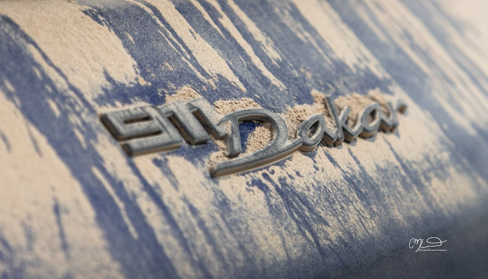 Porsche 911 Dakar Roughroads. Close-up of a car emblem on a blue vehicle, partially covered with sand.