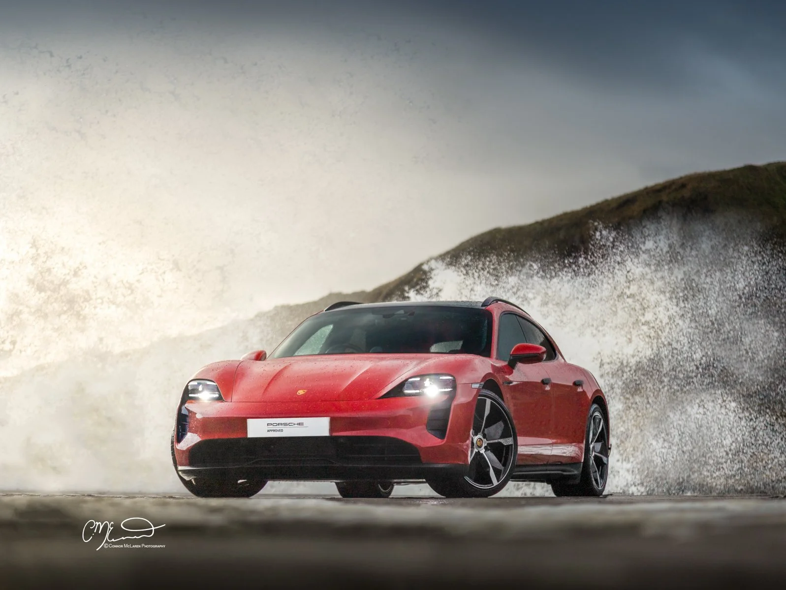 A red Porsche Taycan sports car driving on a wet road with crashing waves and cliffs in the background.