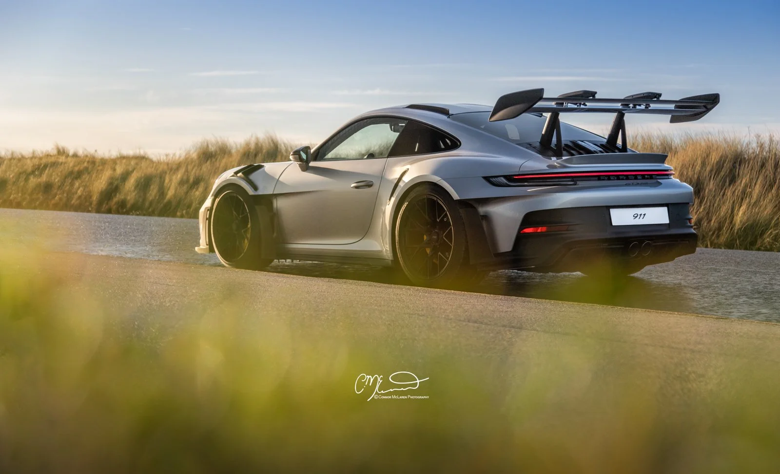 A silver Porsche 911 GT3 RS, 992 sports car with a large rear wing driving on a wet road in a grassy field during sunset.