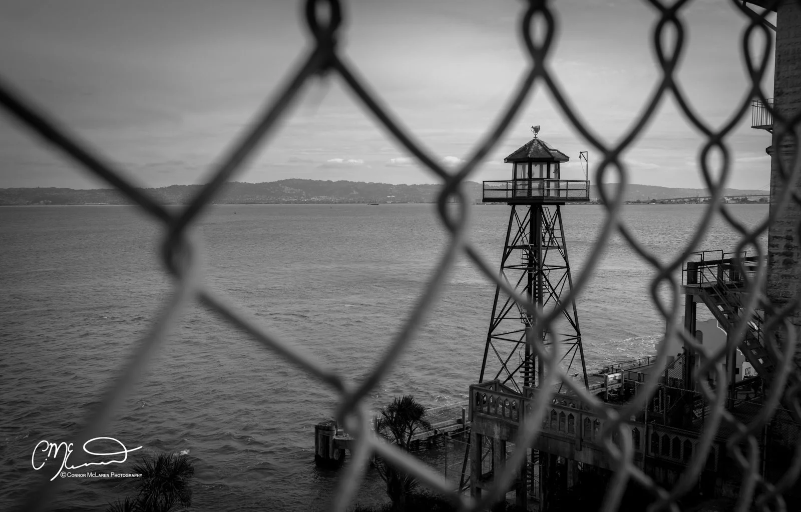 USA Alcatraz California. A black and white photograph of a metal lighthouse structure seen through a chain-link fence, overlooking a body of water with distant hills in the background.
