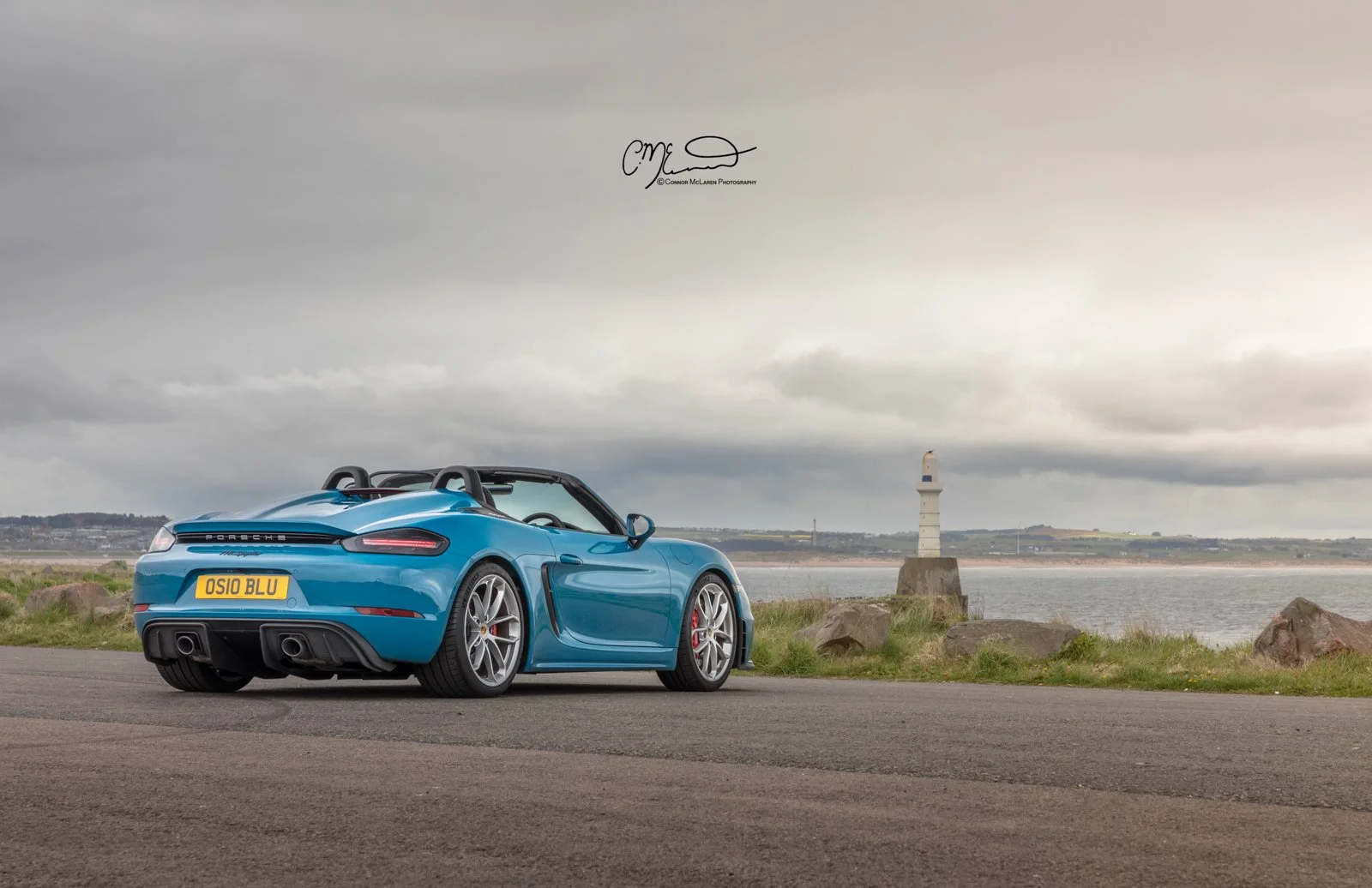 A Oslo Blue Porsche Spyder convertible sports car parked near a body of water with a lighthouse in the background and a cloudy sky overhead.