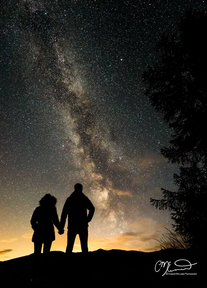 Perthshire, Night Sky, Scotland.  Silhouettes of a man and a woman holding hands and looking at the starry night sky, with the Milky Way galaxy visible overhead.