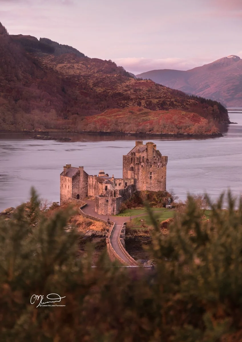 Bucket list locations, Scotland, Eilean Donan Castle. A castle on a small island connected to the mainland by a narrow bridge, with surrounding water and mountains in the background during sunset or sunrise.