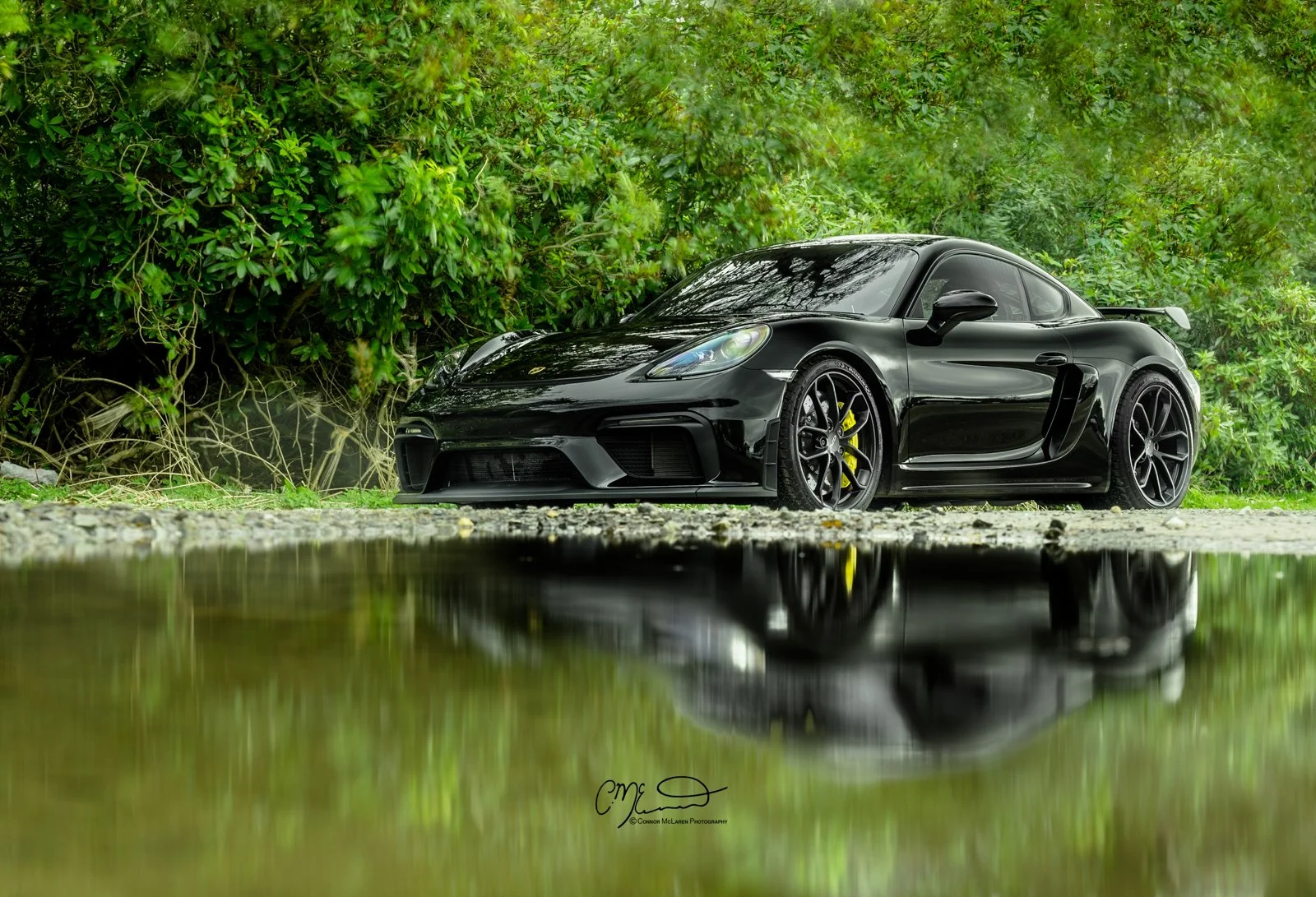 A black Porsche 718 GT4 sports car parked on a gravel surface near a green bush with dense foliage, with a reflection of the car in a puddle of water in the foreground.
