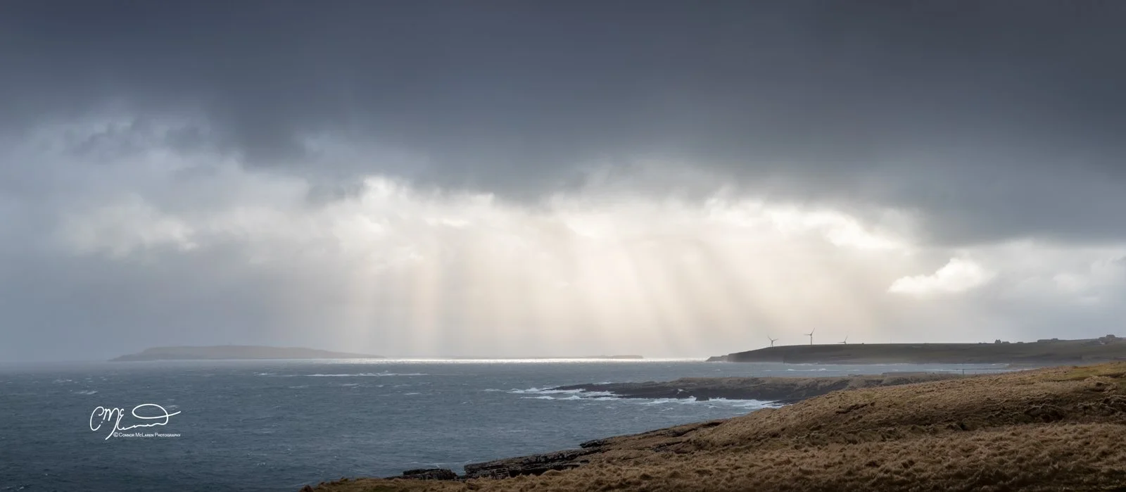 Orkney, Scotland. Overcast sky over a coastal landscape with waves crashing on rocks, wind turbines on a distant hill, and sunbeams breaking through clouds.