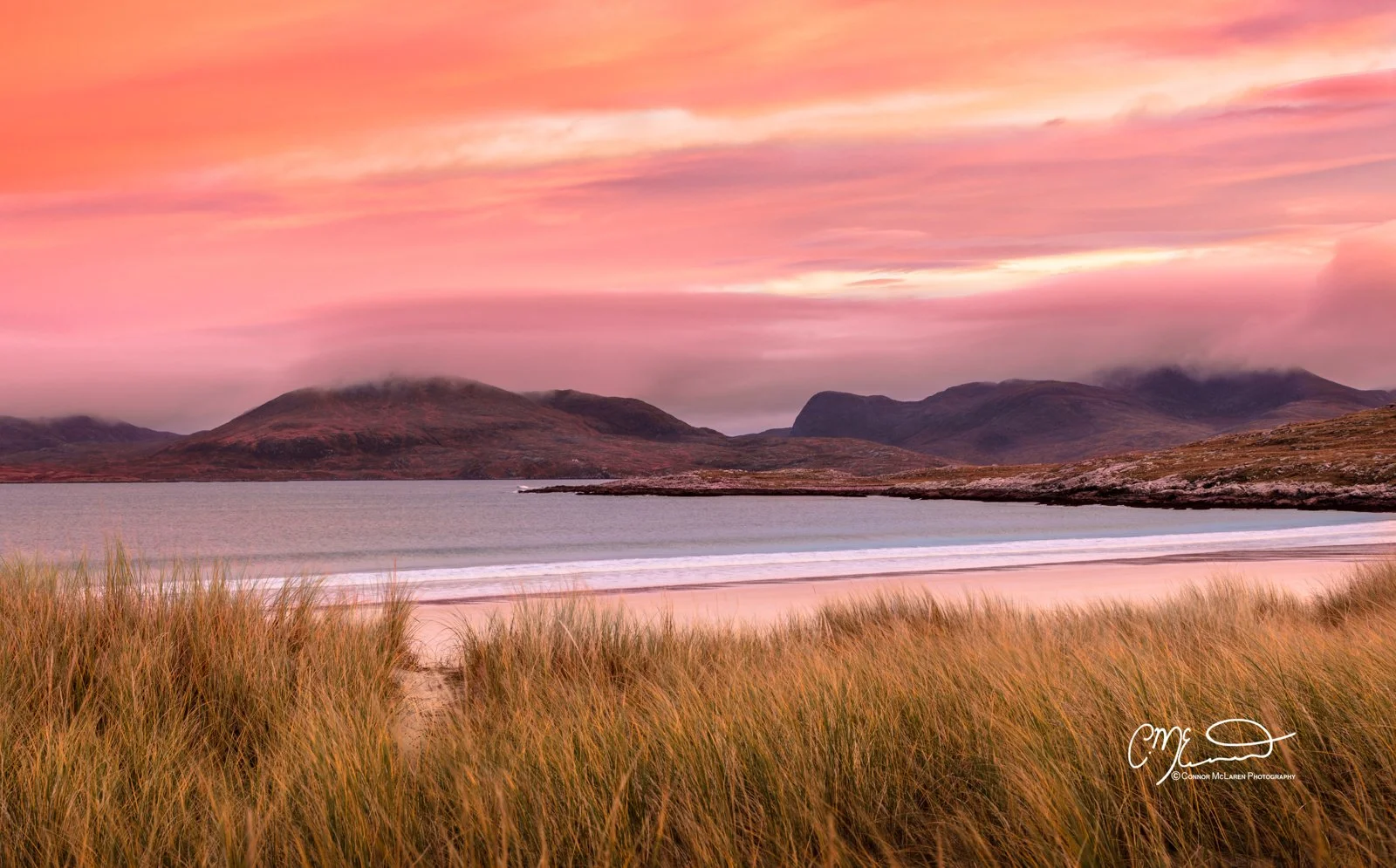 Bucket list locations, Scotland, isle of Harris, Luskentyre Beach. A serene landscape of a beach with tall grasses in the foreground, calm water, and mountains in the background under a pink and orange sunset sky.